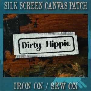 May include: A rectangular off-white canvas patch with frayed edges featuring a black rectangular border and the text "Dirty Hippie" printed in a black script font. The patch is designed for iron-on or sew-on application to fabric surfaces.