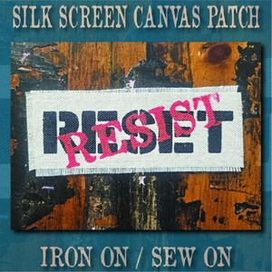 May include: A rectangular off-white canvas patch featuring the word RESIST printed in bold black block letters with the word RESIST also printed in a bright pink script font layered directly over the black text, with frayed edges on the fabric.
