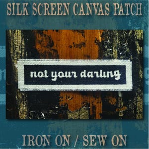 May include: A rectangular canvas patch featuring a black centre with the white text "not your darling" in a lowercase serif font, surrounded by a light-coloured border with frayed edges. The patch is designed for iron-on or sew-on application.