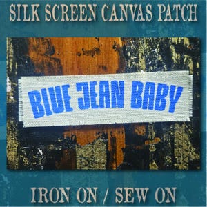 May include: A rectangular canvas patch with frayed edges featuring the text BLUE JEAN BABY printed in a bold blue font. The patch is positioned against a dark, textured background. The text SILK SCREEN CANVAS PATCH and IRON ON / SEW ON appear.
