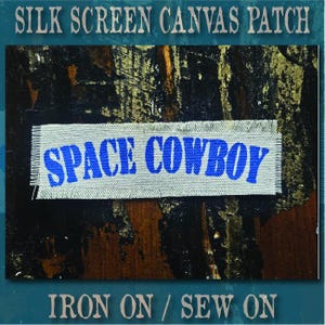 May include: A rectangular canvas patch with frayed edges featuring the text SPACE COWBOY printed in a bold blue font. The patch is light gray in color and has a textured fabric appearance with visible weave patterns throughout the material.