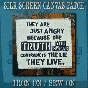 Può includere: Una toppa quadrata in tela color bianco sporco con bordi sfilacciati e testo serigrafato in nero: THEY ARE JUST ANGRY BECAUSE THE TRUTH YOU SPEAK CONTRADICTS THE LIE THEY LIVE. La toppa è mostrata su uno sfondo scuro e strutturato.