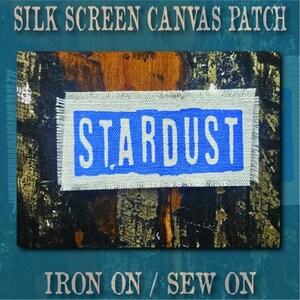 May include: A rectangular canvas patch with frayed edges featuring a blue rectangular background with the word STARDUST printed in white capital letters. The text SILK SCREEN CANVAS PATCH and IRON ON / SEW ON appears above and below the patch.