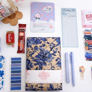 Pu&ograve; includere: Una collezione di articoli a tema librario che include un bicchiere con stampa di libreria, una maschera viso alla pesca, un blocco note per la lista di lettura, un biscotto Biscoff, una bustina di Nescaf&eacute;, una candela blu a forma di libro, un segnalibro floreale, foglietti adesivi, un libro incartato, penne e adesivi.