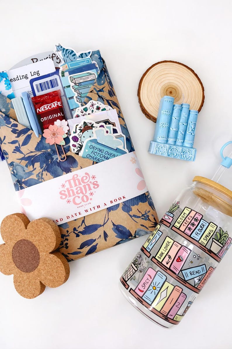 Pu&ograve; includere: Un libro avvolto in carta floreale blu con una fascia The Shan's Co. Book Club, una statuetta azzurra a forma di libri, un sottobicchiere in sughero a forma di fiore e un bicchiere di vetro con coperchio in bamb&ugrave; e stampa di libreria.