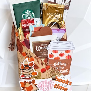 May include: A gift package wrapped in autumn-themed paper with leaf patterns, containing various coffee and hot chocolate packets including Starbucks, Mokate Gold, and Carnation. It features stickers of books and coffee cups with the text Falling Into A Book.