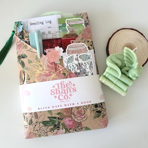 May include: A book club gift set with a floral design. The set includes a decorative envelope with the text "The Shan's Co. Book Club Blind Date With A Book", a reading log, stickers, a pen, and a green dragon candle on a wooden slice.