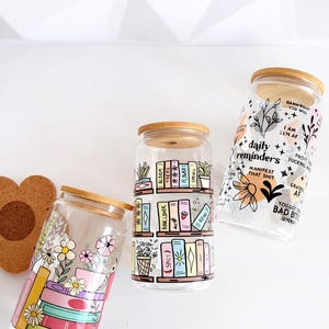 Pu&ograve; includere: Tre bicchieri in vetro trasparente con coperchi in bamb&ugrave;. Il primo presenta libri impilati e fiori. Il secondo mostra scaffali di libri con etichette come Bloom, Trees e Book Lover. Il terzo mostra testo, inclusi daily reminders e manifest that shit.