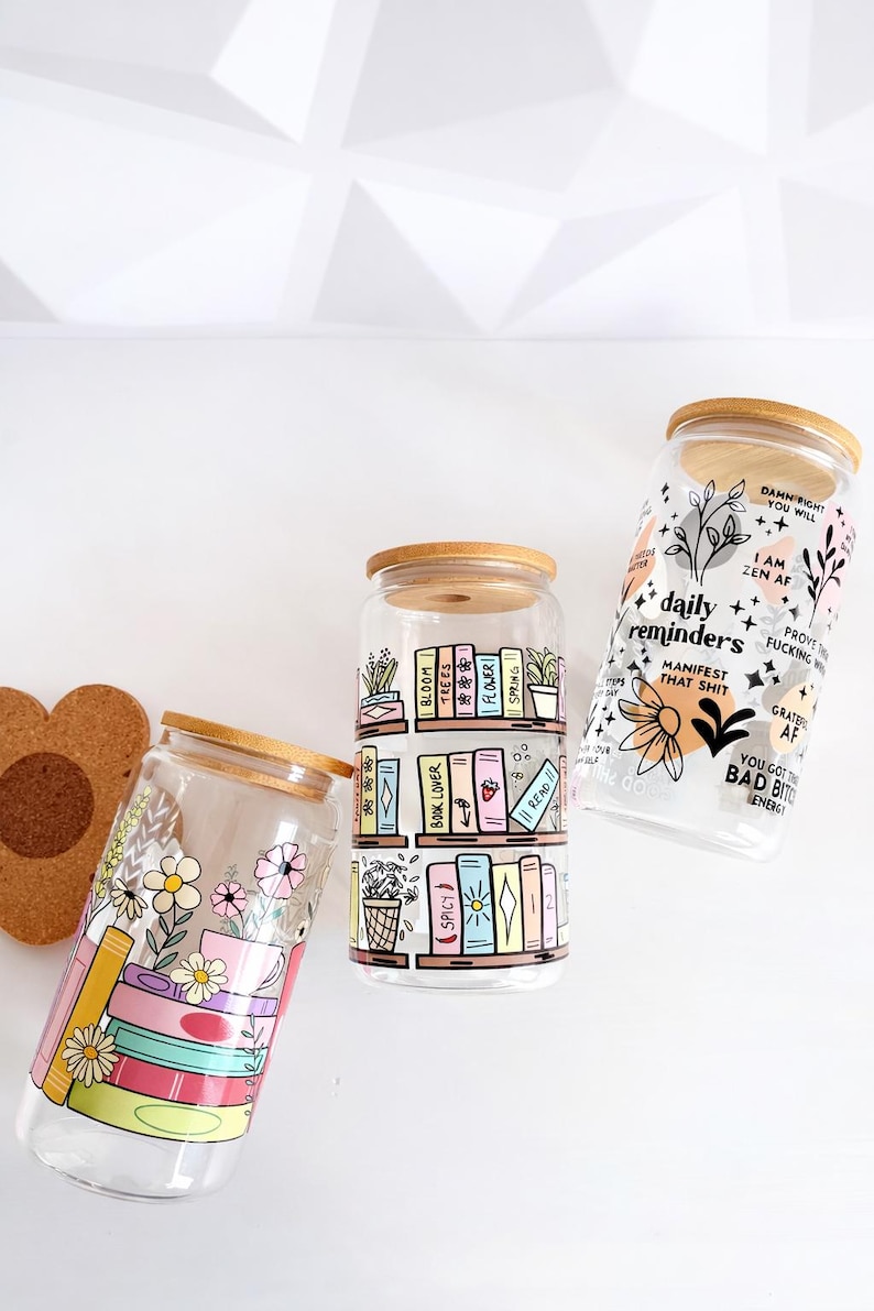 Pu&ograve; includere: Tre bicchieri in vetro trasparente con coperchi in bamb&ugrave;. Il primo presenta libri impilati e fiori. Il secondo mostra scaffali di libri con etichette come Bloom, Trees e Book Lover. Il terzo mostra testo, inclusi daily reminders e manifest that shit.