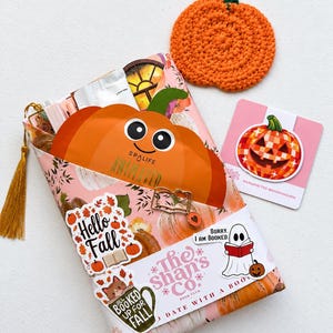 May include: A gift set featuring a pumpkin-shaped face mask, a crocheted orange pumpkin coaster, and a magnetic bookmark with a pumpkin graphic. The package is wrapped in floral paper with stickers reading Hello Fall and All Booked Up For Fall.