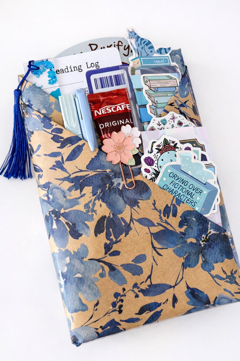 Pu&ograve; includere: Una busta color marrone chiaro con motivo floreale blu contenente un diario di lettura, una nappa blu, due penne azzurre, una bustina di caff&egrave; Nescaf&eacute; Original, una graffetta a forma di fiore rosa e vari adesivi, tra cui uno con scritto Crying Over Fictional Characters.