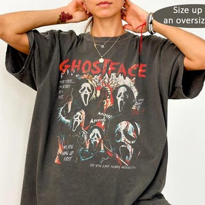 May include: A charcoal grey short-sleeve t-shirt featuring a graphic print of the Ghostface mask from the Scream film franchise. The design includes red text reading GHOSTFACE and various movie quotes. The shirt has a loose, oversized fit.