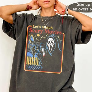 May include: A charcoal gray short-sleeve t-shirt featuring a graphic print with the text Let's watch Scary Movies. The design depicts a hooded figure holding a knife in front of a house, trees, and a fence, framed by a yellow and red border.