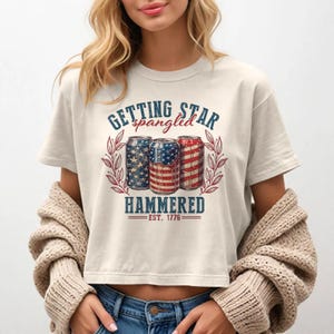 May include: Cream-colored crop top with a graphic featuring three beer cans decorated with American flag designs. The text reads "Getting Star Spangled Hammered" with "Est. 1776" below. The design is framed by a red and green wreath.