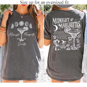 May include: A charcoal gray t-shirt featuring white graphic prints. The front displays moon phases, a margarita glass, and the text Midnight Margarita Society ESTD 1692. The back shows a large margarita glass, a frog, and text.