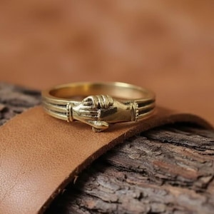 May include: A gold-colored ring featuring a detailed design of two clasped hands. The ring is set against a backdrop of brown leather and textured wood, highlighting its intricate craftsmanship and vintage aesthetic.