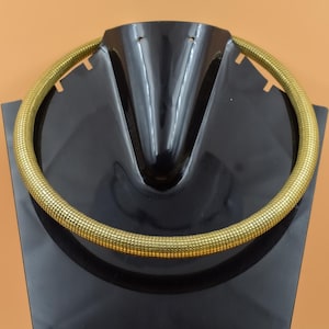 Puede incluir: Un collar dorado y texturizado, exhibido en un expositor de joyería negro. El collar tiene un diseño grueso y redondeado, y la superficie parece estar hecha de pequeños segmentos conectados. El expositor es brillante, contrastando con el acabado mate del collar.