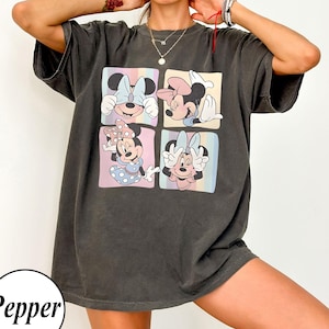 May include: Dark grey t-shirt with a four-panel graphic of Minnie Mouse in different poses. Each panel has a pastel background. The shirt has short sleeves and a relaxed fit. The word "Pepper" is in a white circle at the bottom.
