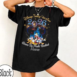 May include: Black t-shirt featuring a colorful graphic of Disney characters in front of a castle, with the text "Disney Family Vacation 2026" and "Where the Magic Begins! Name". The shirt has a round neckline and short sleeves.