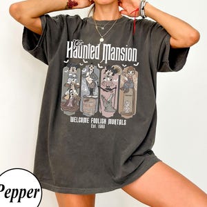 Könnte beinhalten: Dunkelgraues T-Shirt mit einem Motiv von The Haunted Mansion. Das Design enthält den Text "The Haunted Mansion" und "Welcome Foolish Mortals Est. 1969" zusammen mit Cartoon-Figuren. Das T-Shirt ist übergroß.