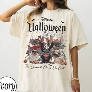 Könnte beinhalten: Elfenbeinfarbenes T-Shirt mit einem Disney Halloween-Motiv. Das Design zeigt Stitch-Charaktere in Halloween-Kostümen, mit dem Text "Disney Halloween" und "The Spookiest Place On Earth". Fledermäuse und Kürbisse sind ebenfalls enthalten.
