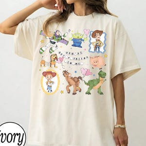 Puede incluir: Camiseta de color marfil con personajes de dibujos animados coloridos de una popular película animada. El diseño incluye a Buzz Lightyear, un extraterrestre, Woody, Jessie, Slinky Dog, Hamm, Bullseye y Rex, con la frase "You've Got a Friend in Me."