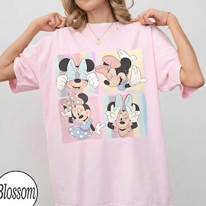 May include: A light pink t-shirt featuring a four-panel graphic of Minnie Mouse in various poses. Each panel has a pastel rainbow background. The shirt has short sleeves and a round neckline. The word "Blossom" is in a circle at the bottom left.
