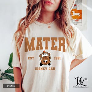 May include: Ivory t-shirt with the word "MATER" in a brown, collegiate font. Below is a cartoon car image and the text "EST 1951 DISNEY CAR". The shirt has a crew neck and short sleeves. The colour of the shirt is ivory.