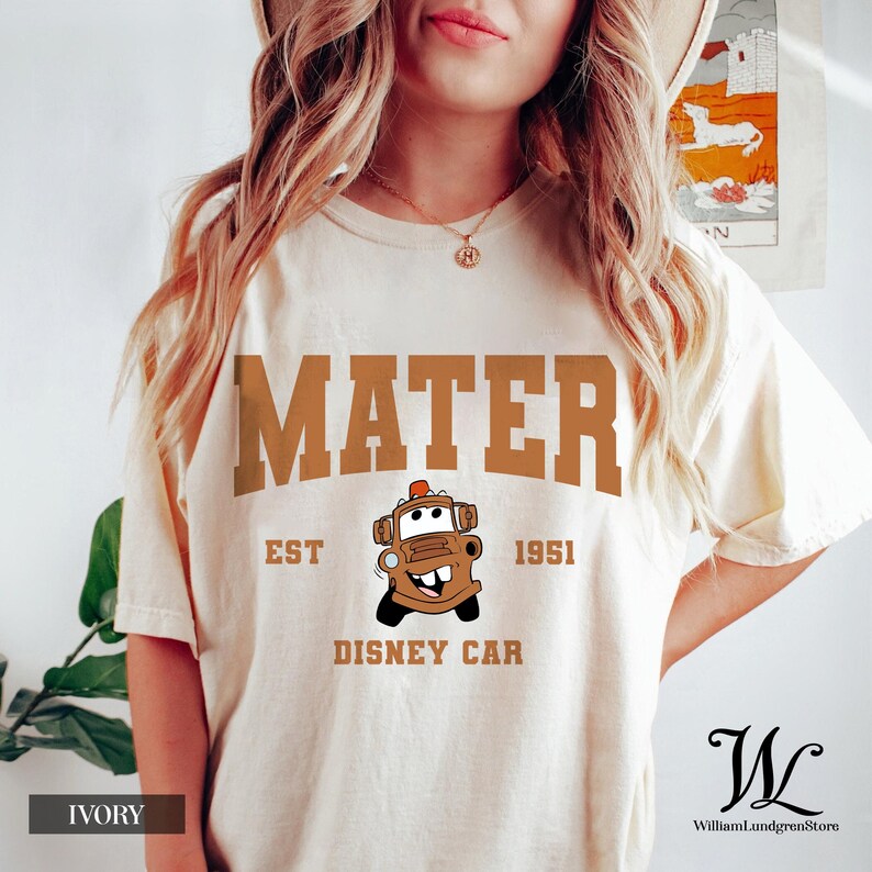 May include: Ivory t-shirt with the word "MATER" in a brown, collegiate font. Below is a cartoon car image and the text "EST 1951 DISNEY CAR". The shirt has a crew neck and short sleeves. The colour of the shirt is ivory.