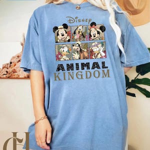 May include: A light blue t-shirt featuring a Disney graphic with Mickey Mouse, Minnie Mouse, Donald Duck, and Goofy. The shirt has the words "Disney Animal Kingdom" printed on the front.