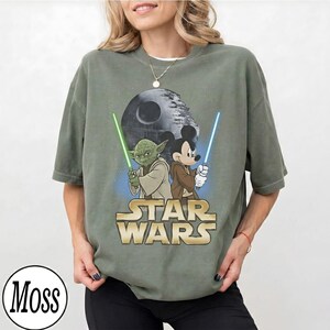 May include: Olive green t-shirt featuring a graphic of Yoda and Mickey Mouse holding lightsabers, with the Death Star in the background. The words "STAR WARS" are printed in gold. The shirt is a relaxed fit.