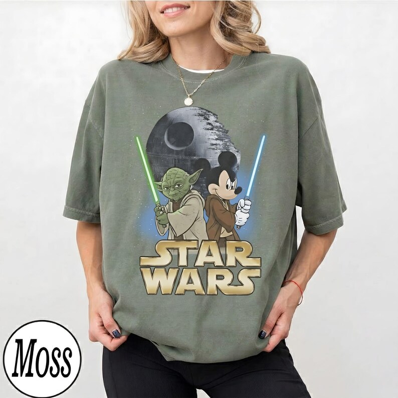 May include: Olive green t-shirt featuring a graphic of Yoda and Mickey Mouse holding lightsabers, with the Death Star in the background. The words "STAR WARS" are printed in gold. The shirt is a relaxed fit.
