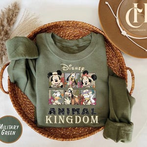 May include: Military green sweatshirt featuring Disney characters in a grid design. The sweatshirt has the Disney logo and the words "Animal Kingdom" in a bold font. The sweatshirt is displayed in a woven basket.