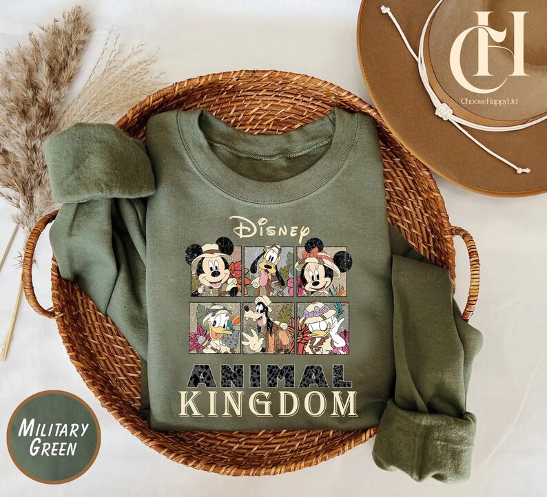 May include: Military green sweatshirt featuring Disney characters in a grid design. The sweatshirt has the Disney logo and the words "Animal Kingdom" in a bold font. The sweatshirt is displayed in a woven basket.
