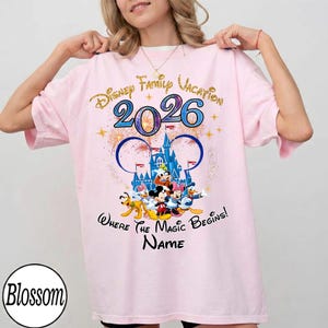 May include: Light pink t-shirt with a Disney Family Vacation 2026 design. The design features the Disney castle, fireworks, and Disney characters. The text "Where the Magic Begins!" and a space for a name are also included.