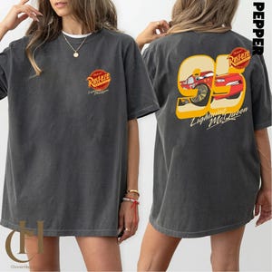 以下が含まれることがあります： 前面に「Rusteze」のロゴ、背面に95番のライトニング・マックィーンのグラフィックがプリントされたダークグレーのTシャツ。「Lightning McQueen」の文字が含まれています。