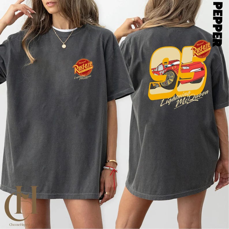 以下が含まれることがあります： 前面に「Rusteze」のロゴ、背面に95番のライトニング・マックィーンのグラフィックがプリントされたダークグレーのTシャツ。「Lightning McQueen」の文字が含まれています。