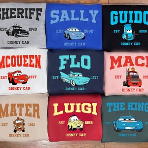 May include: Nine sweatshirts featuring Disney Cars characters. Each sweatshirt displays a character name, a car illustration, and the text "Disney Car". The sweatshirts are in various colours, including grey, blue, black, white, navy, pink, tan, red, and teal.