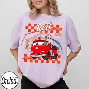 May include: A light purple t-shirt featuring a cartoon fire truck with the word "Red" and the year "2006" in orange and white. The fire truck is red with blue water spraying from the hose. The shirt has a checkered orange and white pattern.