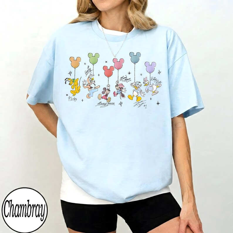 May include: Light blue t-shirt featuring a colorful graphic of Disney characters holding Mickey Mouse-shaped balloons. The design includes Pluto, Donald Duck, Mickey Mouse, and others. The shirt has short sleeves and a relaxed fit.