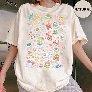 Puede incluir: Camiseta color crema con un colorido gráfico de Super Mario. El diseño incluye varios personajes y elementos del juego, como Mario, champiñones, estrellas y mariposas. Las palabras "Super Mario" están escritas en cursiva.