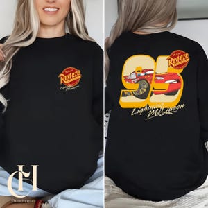 以下が含まれることがあります： 「Rust-eze」のロゴと「Lightning McQueen」のグラフィックが特徴の黒いスウェットシャツ。前面にはロゴ、背面にはLightning McQueenのイラストが入った大きな「95」が表示されています。デザインは赤、黄、白です。