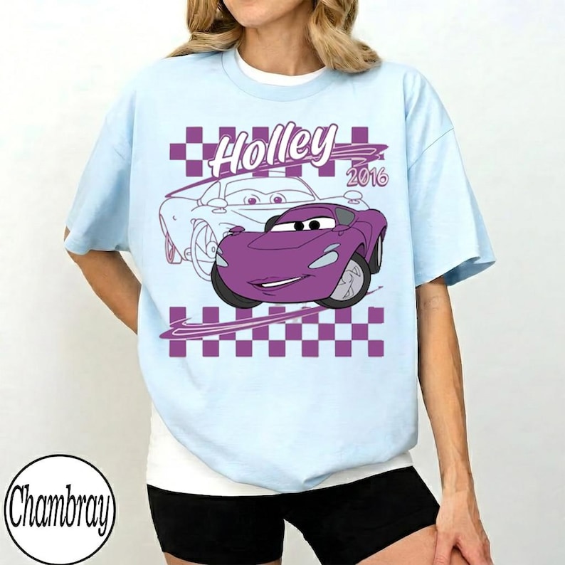 May include: Light blue t-shirt featuring a purple cartoon car with the name "Holley" and the year "2016" printed on the front. The design includes a checkered pattern and a light blue outline of a car. The shirt has short sleeves.