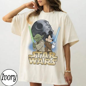 May include: Ivory-colored t-shirt featuring a Star Wars graphic with Yoda and Mickey Mouse holding lightsabers. The Star Wars logo is in gold. The Death Star is in the background.