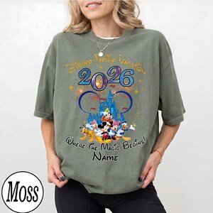 May include: Olive green t-shirt featuring a Disney Family Vacation design for 2026. The graphic includes Disney characters, a castle, fireworks, and the text "Where the Magic Begins!" and "Name".