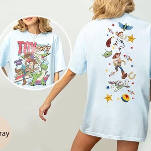 May include: A light blue t-shirt featuring Toy Story graphics. The front displays Woody and Rex with the text "Toy Story". The back features a vertical arrangement of characters including Woody, Buzz Lightyear, an alien, and various icons like stars, rockets, and a ball.