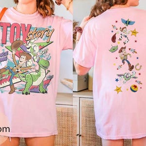 May include: A light pink t-shirt featuring Toy Story graphics. The front displays Woody, Buzz Lightyear, and Rex with the text "TOY story SINCE 1995". The back features a vertical arrangement of various characters and icons including Woody, Buzz, and an alien.