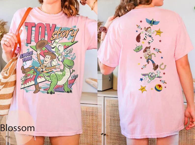 May include: A light pink t-shirt featuring Toy Story graphics. The front displays Woody, Buzz Lightyear, and Rex with the text "TOY story SINCE 1995". The back features a vertical arrangement of various characters and icons including Woody, Buzz, and an alien.