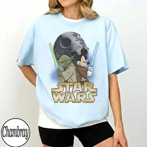 May include: Light blue t-shirt featuring a graphic of Yoda and Mickey Mouse holding lightsabers, with the Death Star in the background. The Star Wars logo is displayed in gold. The shirt is a relaxed fit.