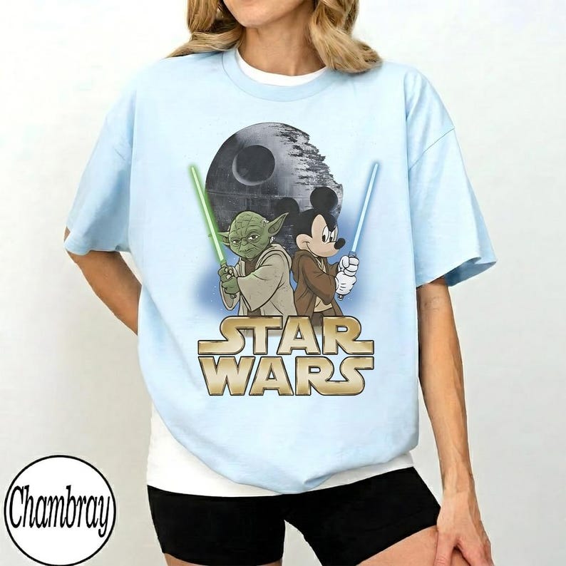 May include: Light blue t-shirt featuring a graphic of Yoda and Mickey Mouse holding lightsabers, with the Death Star in the background. The Star Wars logo is displayed in gold. The shirt is a relaxed fit.